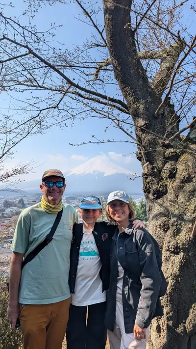 The three of us posed against a big cherry trunk on a hillside above town, Mount Fuji snow-capped and clear in the gap behind us. Charlie in the middle wearing a white t-shirt that reads "Mt. Fuji 3,776" with the mountain itself directly over his shoulder. Bright, hazy, blue sky; the tree is still bare buds, no blossom out yet at this altitude.