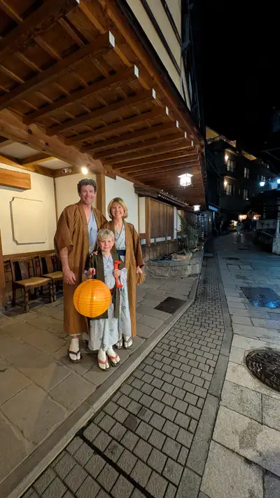 All three of us on the stone street in Shibu Onsen at night, in the ryokan yukata with striped haori over them and geta with white tabi socks. Charlie in front holding a lit orange paper lantern and a red something in his other hand. Wet cobbles, wooden eaves overhead, hanging lanterns and a few other guests further up the street.